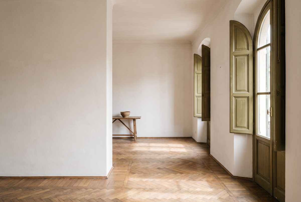 Soggiorno vuoto e luminoso di un appartamento in affitto, con parquet a spina di pesce e persiane verde oliva aperte