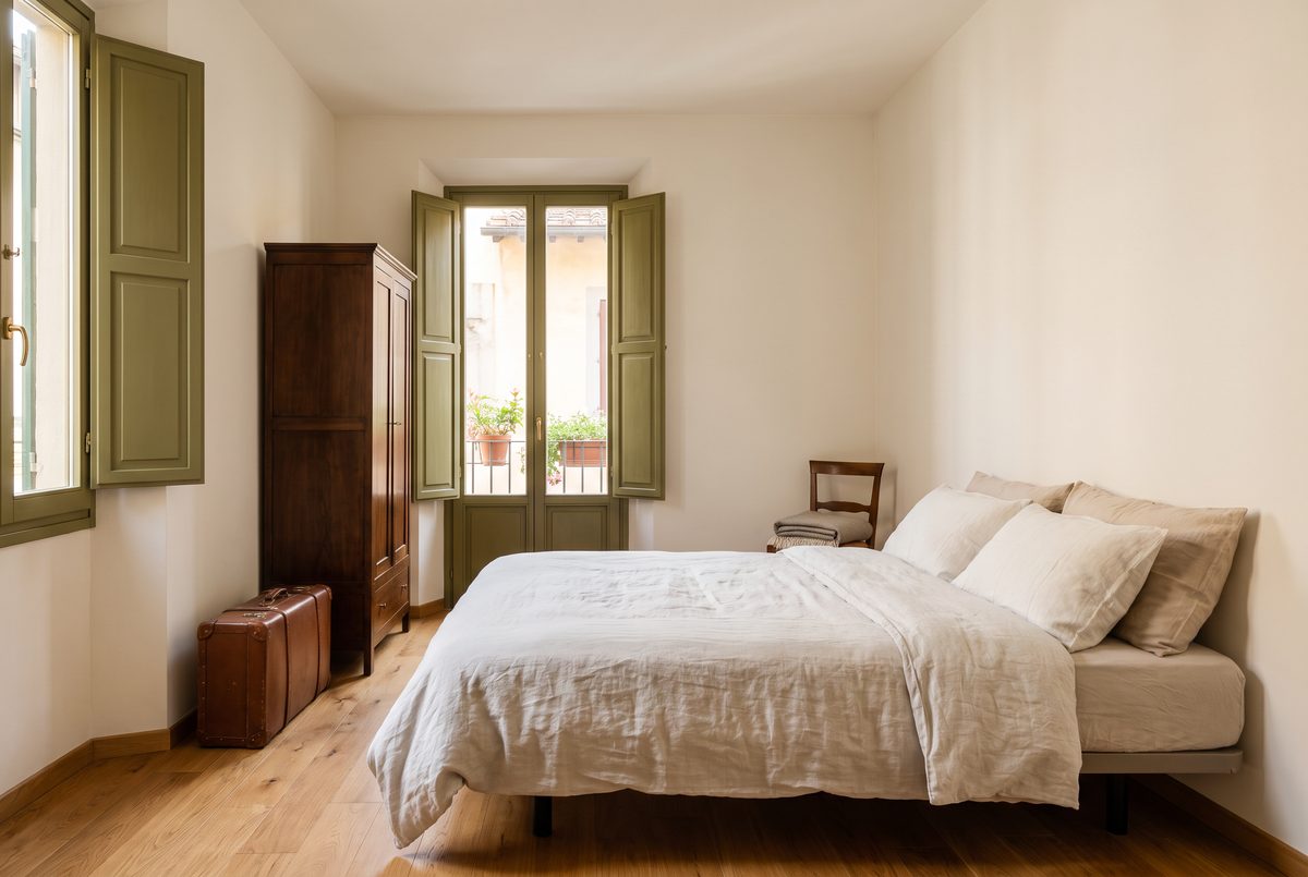 Camera arredata e luminosa con parquet, letto rifatto, una valigia chiusa accanto all'armadio e persiane verde oliva socchiuse