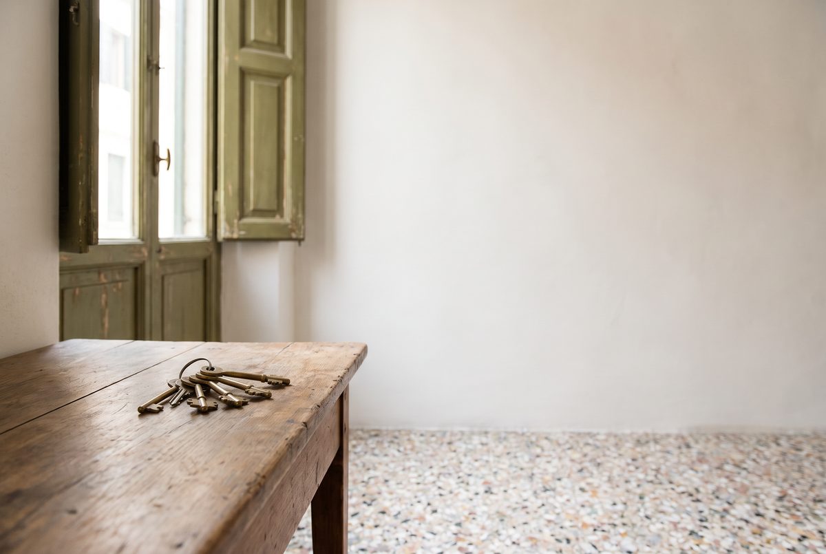 Mazzo di vecchie chiavi di ottone su un tavolo di legno, accanto a una finestra con persiane verde oliva e a un pavimento in graniglia