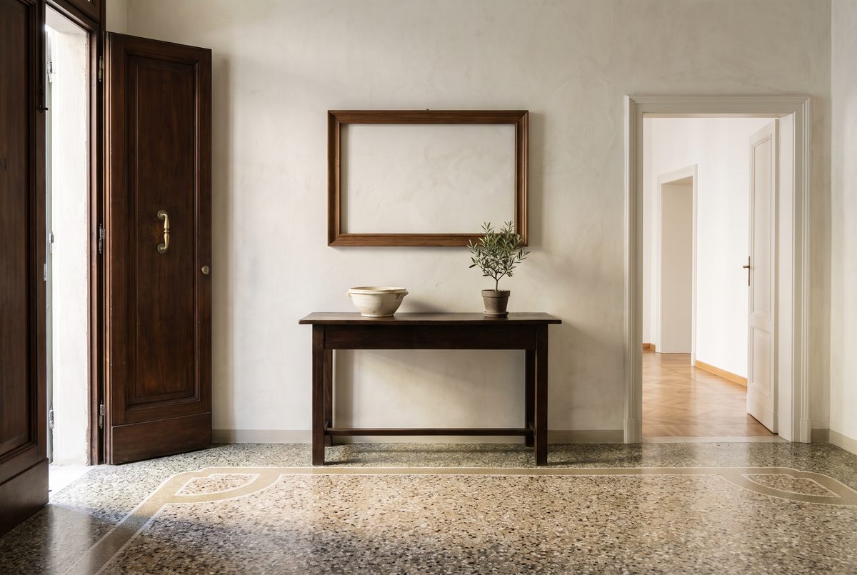 Ingresso di un appartamento con pavimento in graniglia, una consolle di legno con una ciotola di ceramica e una piccola pianta, luce laterale del mattino